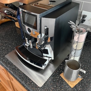 May include: A white ceramic mug with a black handle and the text "But First, Coffee" in a bold, black font. The mug is filled with a dark liquid, likely coffee, and sits on a light-colored surface.