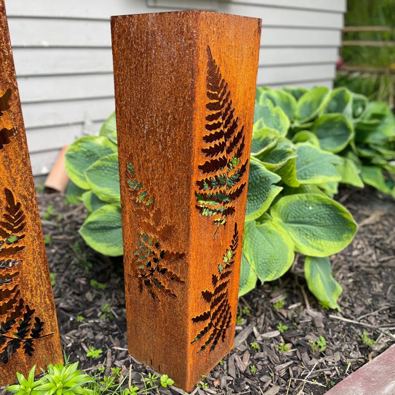 May include: Two rusty metal garden stakes with fern cutouts. The stakes are standing upright in a garden setting.
