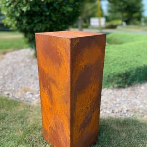 May include: A tall, rectangular planter with a weathered, rusty orange finish. The planter has a square top and a slightly tapered shape, sitting on a grassy surface. The outdoor decor piece is made of metal.