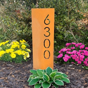 May include: A rusty metal house number sign with the numbers 6300 cut out. The sign is standing in a garden with green plants and pink and yellow flowers.