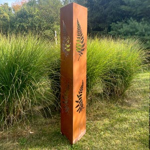 May include: A tall, rectangular, weathered metal garden sculpture. The sculpture has cut-out fern leaf designs and a rusty brown color. It stands in a grassy area with green plants in the background.