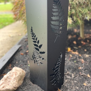 May include: A black metal post with fern leaf cutouts. The post is square and stands upright, with the fern designs visible on multiple sides. The metal has a slight sheen, and the background shows a blurred outdoor setting.