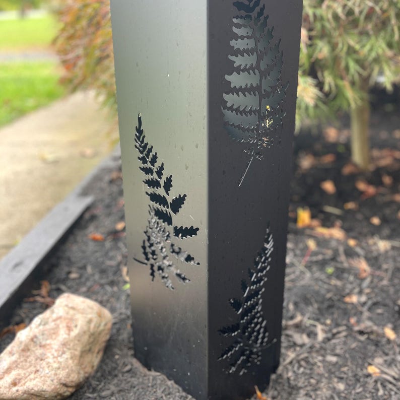 May include: A black metal post with fern leaf cutouts. The post is square and stands upright, with the fern designs visible on multiple sides. The metal has a slight sheen, and the background shows a blurred outdoor setting.