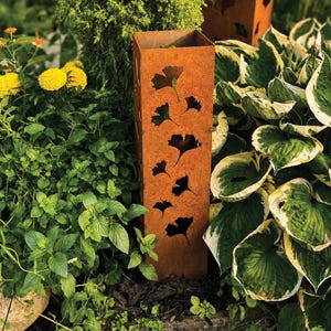 May include: A tall, square, rust-colored metal garden pillar featuring laser-cut ginkgo leaf silhouettes. The vertical column has a weathered, oxidized finish and is positioned upright among various green garden plants and yellow flowers.