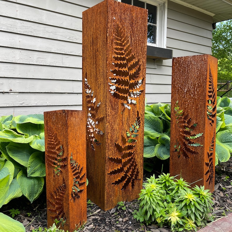 May include: Three tall, rusty metal planters with fern cutouts. The planters are arranged in a row on a bed of mulch and surrounded by green plants.