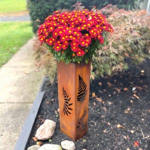 May include: A tall, rectangular, rusted metal planter with fern cutouts, holding a vibrant bouquet of red and yellow chrysanthemums. The planter is outdoors, set in a bed of dark mulch and small rocks.
