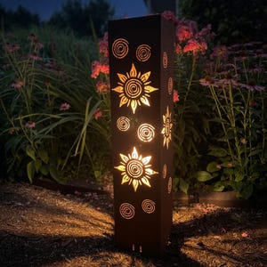 May include: A tall, rectangular, dark metal garden lantern featuring laser-cut designs of suns and spirals. The interior light shines through the cutouts, casting a warm glow against the dark exterior surface of the vertical pillar structure.