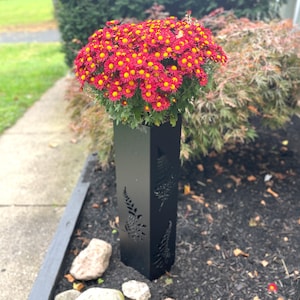 May include: A black metal planter with fern cutouts holds a large cluster of red and yellow chrysanthemums. The planter is rectangular and stands upright in a garden bed with mulch and rocks. The flowers are in full bloom.