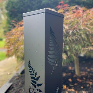 May include: A black metal outdoor post with a square shape and a flat top. The post features cut-out fern leaf designs on its sides. The background shows a blurred view of a garden with trees and foliage.