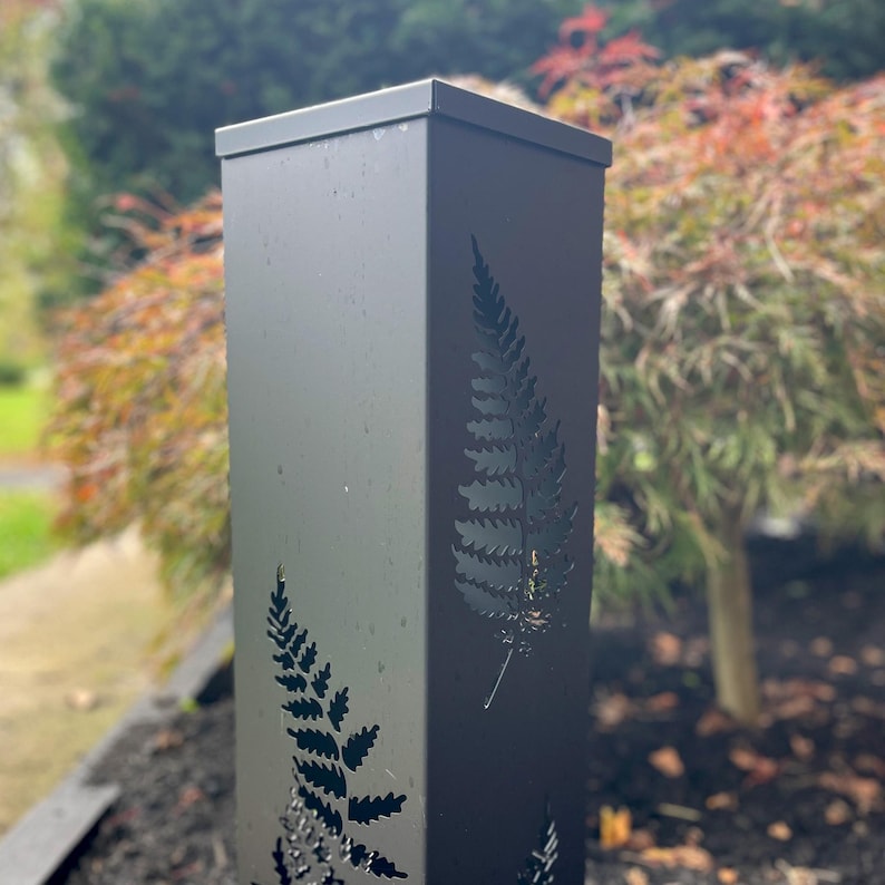 May include: A black metal outdoor post with a square shape and a flat top. The post features cut-out fern leaf designs on its sides. The background shows a blurred view of a garden with trees and foliage.