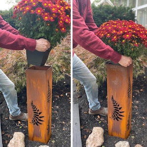 May include: A rectangular, weathered metal planter with fern cutouts. The planter is being used to hold a potted plant with red and yellow flowers. The planter has a rustic, aged appearance, and is outdoors.