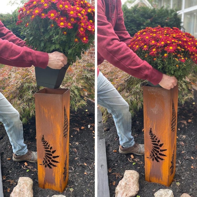May include: A rectangular, weathered metal planter with fern cutouts. The planter is being used to hold a potted plant with red and yellow flowers. The planter has a rustic, aged appearance, and is outdoors.
