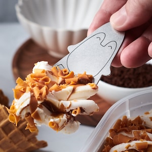 May include: A close-up of a metal ice cream scoop shaped like a penguin with a smiling face. The scoop is holding a scoop of vanilla ice cream with caramel sauce and caramel shavings.