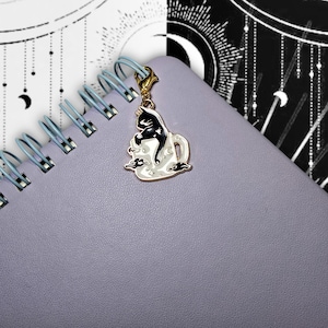 May include: A gold-toned charm with a black cat design inside a white teacup with cloud accents. The charm is attached to a light purple spiral-bound notebook. The background includes a black and white celestial design.