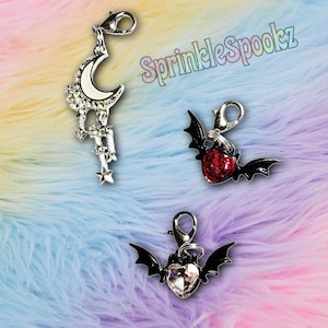 May include: Three decorative charms with lobster clasps. One charm features a crescent moon with rhinestones, another a red heart with bat wings, and the third a clear heart with bat wings. The background is a colorful, fluffy fabric.