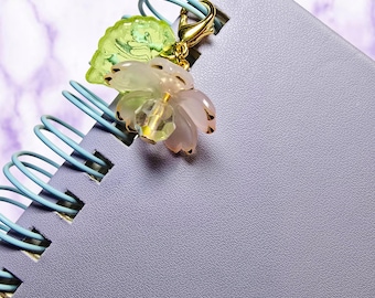 May include: A close-up of a light purple spiral notebook with a light blue spiral binding. A gold-colored charm with a flower and leaf design is attached to the notebook. The flower is pink and clear with gold accents.