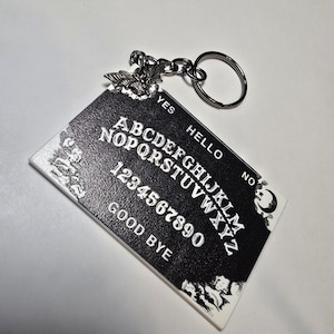 May include: A black and white Ouija board keychain with a silver bat charm. The board features the alphabet, numbers, and the words "YES", "NO", "HELLO", and "GOOD BYE". The keychain has a silver key ring.