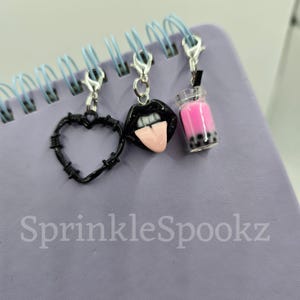 May include: Three black charm pendants with silver clasps. The charms are a heart with barbed wire, a pair of lips with a tongue sticking out, and a clear glass bottle filled with pink liquid and black boba.