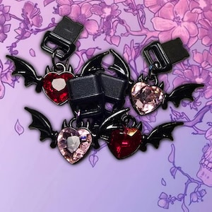 May include: Black metal charms with heart-shaped gems in red and pink, adorned with bat wings. The charms are attached to black clasps, set against a purple floral background. The gems have a faceted cut.