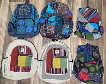 May include: Six backpacks arranged in two rows. The top row features a blue spiral-patterned bag, a multi-colored patchwork bag, and a striped patchwork bag. The bottom row includes two beige bags with colorful striped panels and a dark patchwork bag.