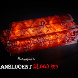 May include: A translucent, blood-red coffin-shaped object with detailed ornamentation. The object is lit, highlighting its intricate design and texture. The image includes the text "Translucent Blood Red" and a logo.