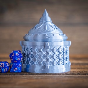 May include: A silver metal dice tower with a detailed design. The tower has a domed top and a base with a chainmail pattern. Three blue dice with gold numbers are in front of the tower.