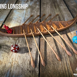 May include: A brown miniature Viking longship model with multiple oars extending outward, featuring a small red-cloaked figure inside. The text VIKING LONGSHIP appears at the top left, and a green, red, and two blue polyhedral dice are nearby.