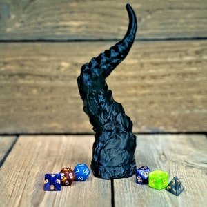 May include: A black 3D printed dragon tail with a textured surface. The tail is standing on a wooden surface with several colorful dice in front of it.