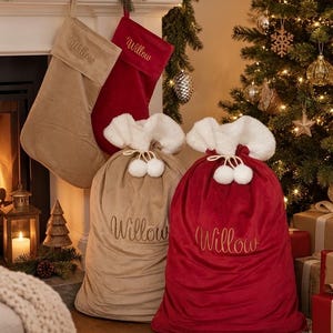 May include: Two velvet Christmas stockings and two matching Santa sacks in beige and red. Each item features the name Willow embroidered in script. The sacks have a white faux fur trim at the top and a drawstring closure with two white pom-poms.