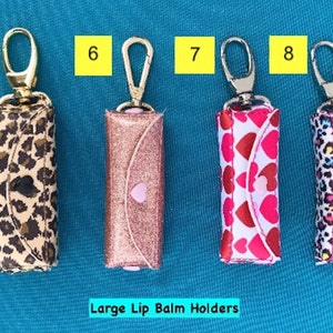 May include: Five different lip balm holders with gold keychains. The holders are made of fabric and have different patterns, including leopard print, glitter, and hearts. The text "Large Lip Balm Holders" is printed below the holders.