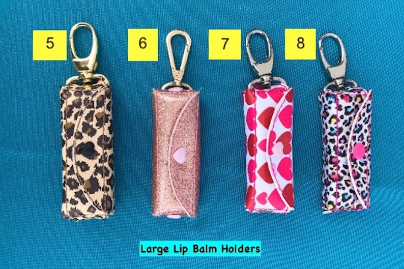 May include: Five different lip balm holders with gold keychains. The holders are made of fabric and have different patterns, including leopard print, glitter, and hearts. The text "Large Lip Balm Holders" is printed below the holders.