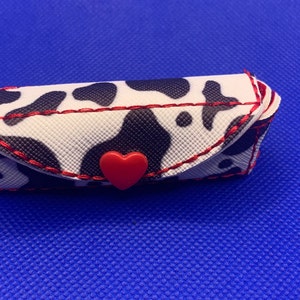 May include: A small, white and black cow print keychain with a red heart-shaped button. The keychain is made of fabric and has a red stitched border.