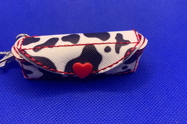 May include: A small, white and black cow print keychain with a red heart-shaped button. The keychain is made of fabric and has a red stitched border.