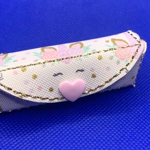 May include: A small white zippered pouch with a pink heart button. The pouch has a gold trim and a unicorn print with pink and green flowers.