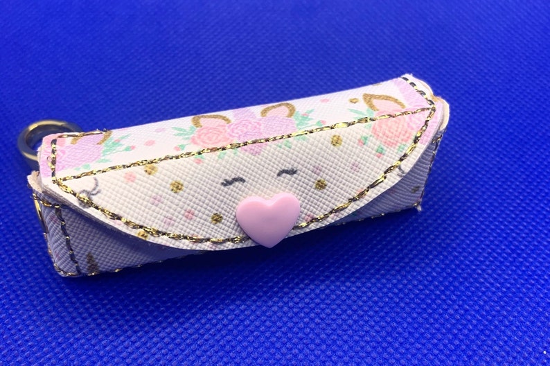 May include: A small white zippered pouch with a pink heart button. The pouch has a gold trim and a unicorn print with pink and green flowers.