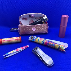 May include: A pink glitter zippered pouch with a lighter and a pink lip balm inside.  The pouch is open and the inside is lined with a white fabric with a pink and white floral pattern.  There is also a small pink and white patterned tube, a silver nail clipper, and a silver tweezers with a white and pink patterned handle on a blue surface.