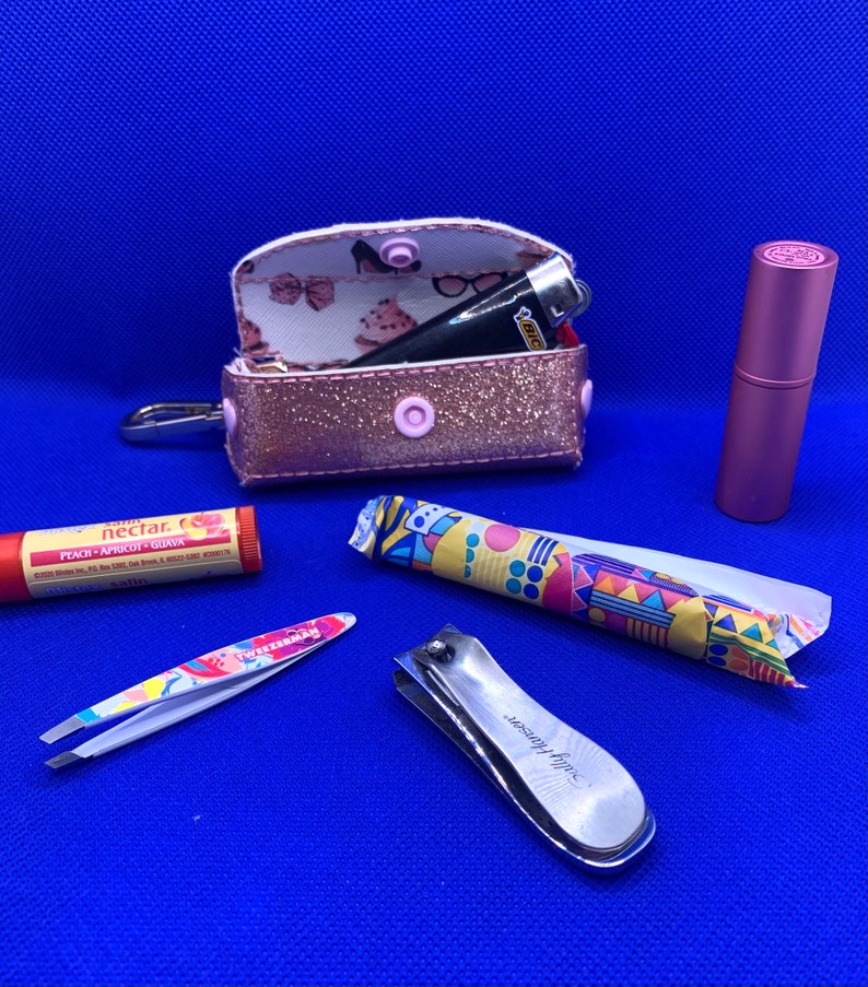 May include: A pink glitter zippered pouch with a lighter and a pink lip balm inside.  The pouch is open and the inside is lined with a white fabric with a pink and white floral pattern.  There is also a small pink and white patterned tube, a silver nail clipper, and a silver tweezers with a white and pink patterned handle on a blue surface.