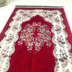 May include: A red prayer rug with a white floral design and a decorative arch. The rug has a fringed edge.