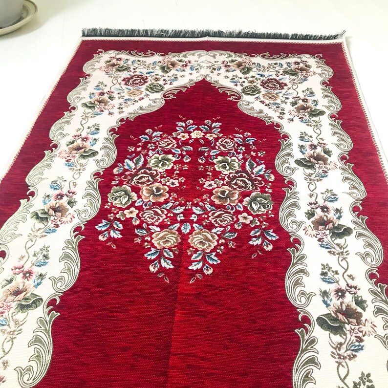 May include: A red prayer rug with a white floral design and a decorative arch. The rug has a fringed edge.