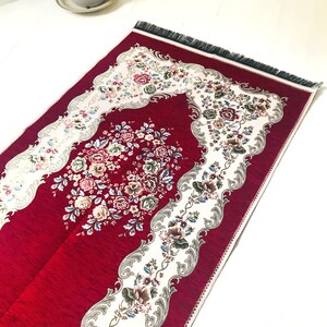 May include: A red prayer rug with a white floral design and fringe. The rug has a traditional Islamic design with a central floral motif.