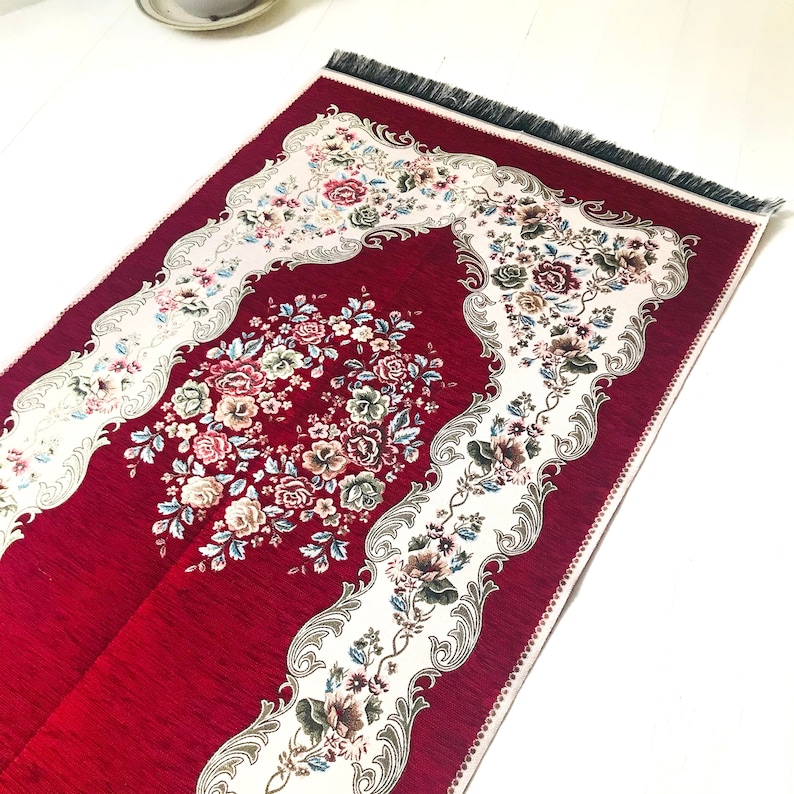 May include: A red prayer rug with a white floral design and fringe. The rug has a traditional Islamic design with a central floral motif.