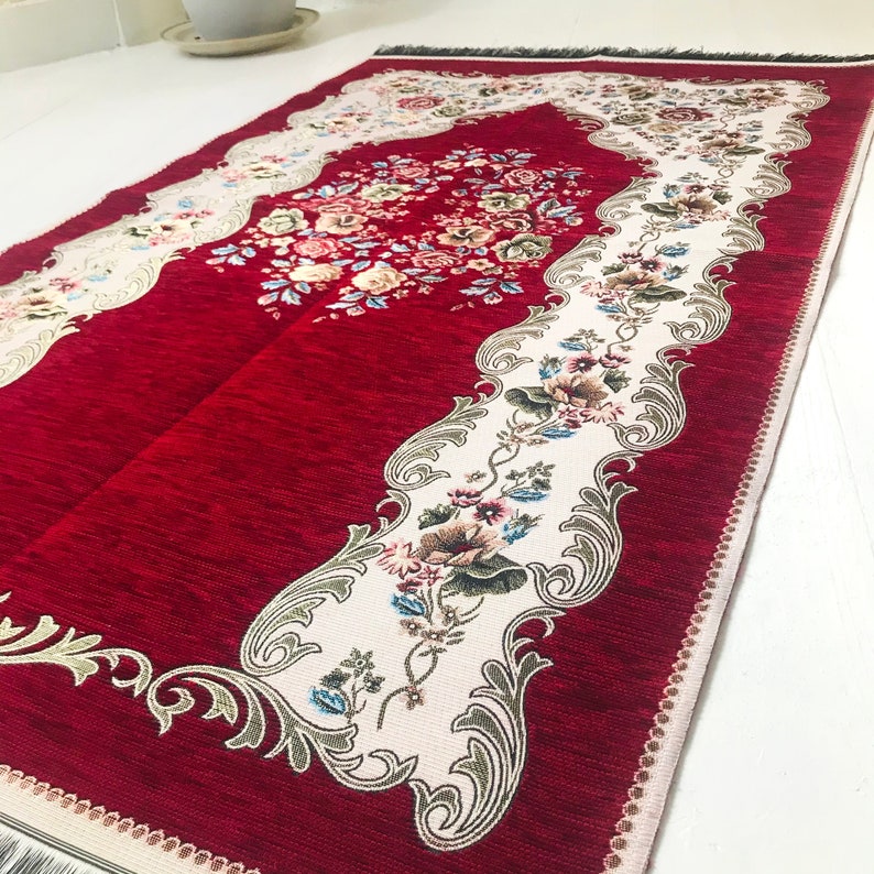 May include: Red prayer rug with a white floral design and a decorative border. The rug has a fringed edge.