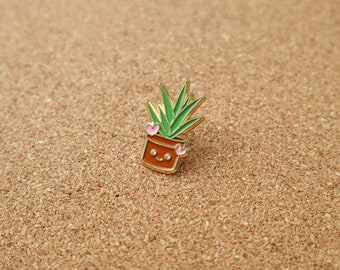 Puede incluir: Un pin de esmalte dorado que representa una planta suculenta verde con corazones rosas para los ojos, plantada en una maceta marrón con una cara sonriente.