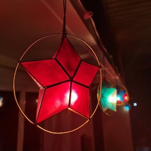 Puede incluir: Una linterna de papel roja en forma de estrella con un marco de alambre dorado cuelga del techo. La linterna está iluminada desde adentro, proyectando un brillo cálido.