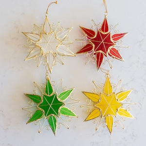Puede incluir: Cuatro adornos de estrellas con marco de alambre con alambre dorado y paneles de vidrio de colores. Las estrellas son blancas, rojas, verdes y amarillas.