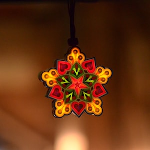 May include: A colorful, star-shaped pendant with a black cord. The pendant features a vibrant design with red hearts, green leaves, and yellow accents. The intricate details and warm colors create a festive and eye-catching piece.