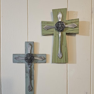 Könnte beinhalten: Zwei dekorative Kreuze aus lackiertem Holz und Besteck. Ein Kreuz ist hellgrün, das andere hellblau lackiert. Jedes Kreuz hat eine dunkle, kreisförmige Mitte und silberne Gabel- und Löffeldetails.