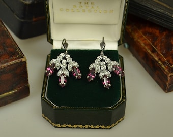 May include: A pair of silver-toned drop earrings featuring marquise-cut deep pink stones and small clear round stones arranged in a floral-inspired cluster design. The earrings are displayed inside a dark green velvet-lined jewelry box.