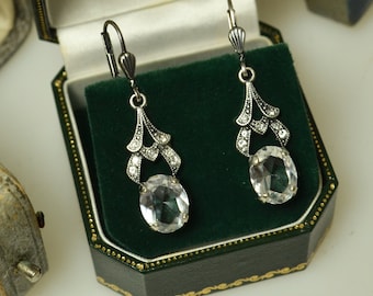 May include: Silver-tone drop earrings with clear oval gemstones. The earrings have an Art Deco design with small, sparkling details. They are presented in a green velvet-lined jewellery box with a black exterior and gold trim.