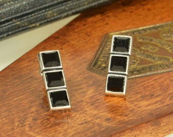 May include: A pair of silver-toned earrings, each featuring three vertically aligned square black stones set in a metallic frame. The earrings have a clean, geometric design with a polished finish, showcasing a linear arrangement of the dark square gems.